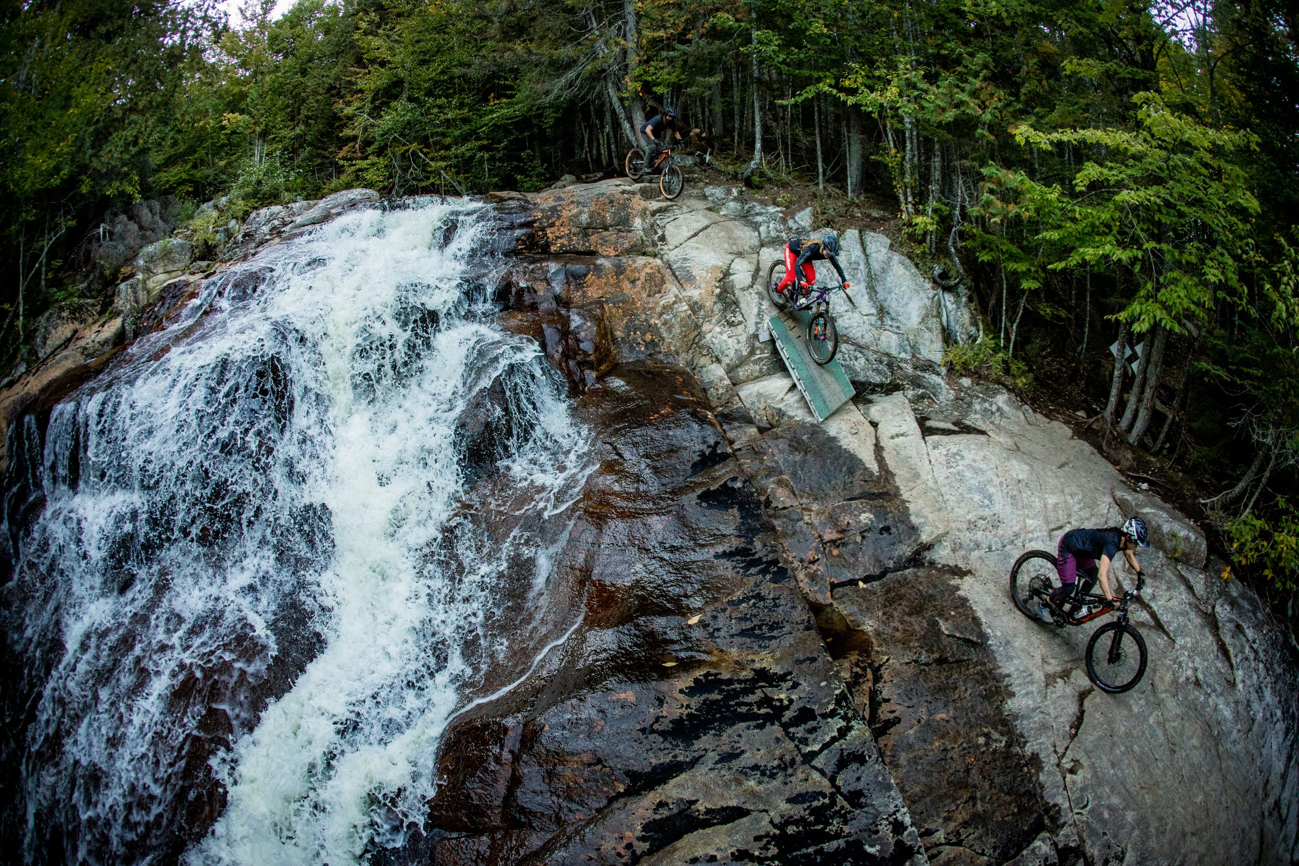 Two cyclists enjoying the outdoor sport of mountain biking on the Légende trail, near a waterfall and on rocky terrain.