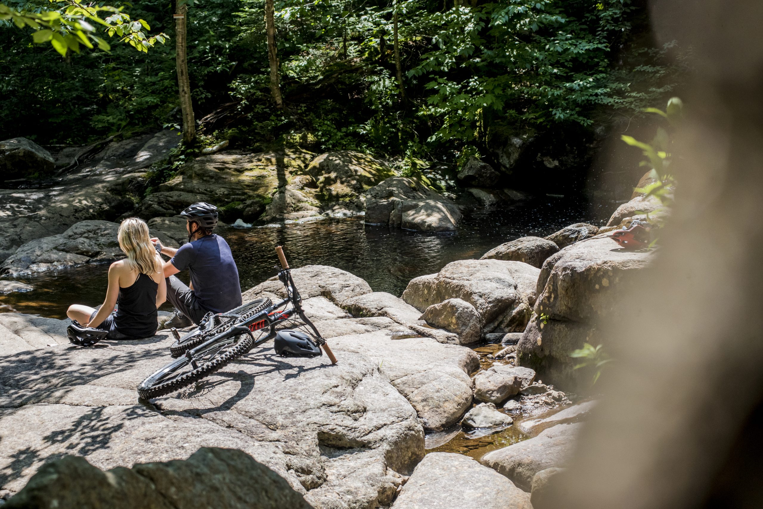 Two cyclists taking a break with their mountain bikes in front of a stream, surrounded by nature, during their outdoor activity.
