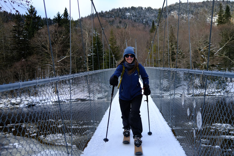 Personne souriante pratiquant le ski-raquette, sur un pont suspendu recouvert de neige blanche, au-dessus de la rivière, avec en arrière-plan les montagnes et la nature hivernale.