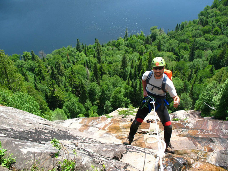 A smiling man in the foreground, indulging in the outdoor activity of Canyoning, on one of the accessible mountains, with a lake in the background and nature all the way down the mountain.