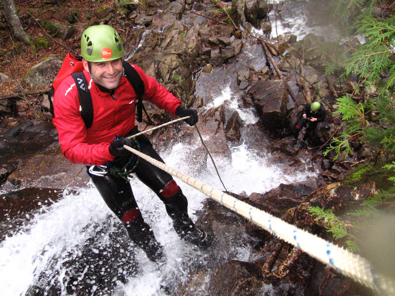 Groupe de personnes, dont un homme souriant en avant-plan, s'adonnant à l'activité de plein air du Canyoning, sur l'une des montagnes accessibles, avec une chute en arrière-plan et la nature à proximité.