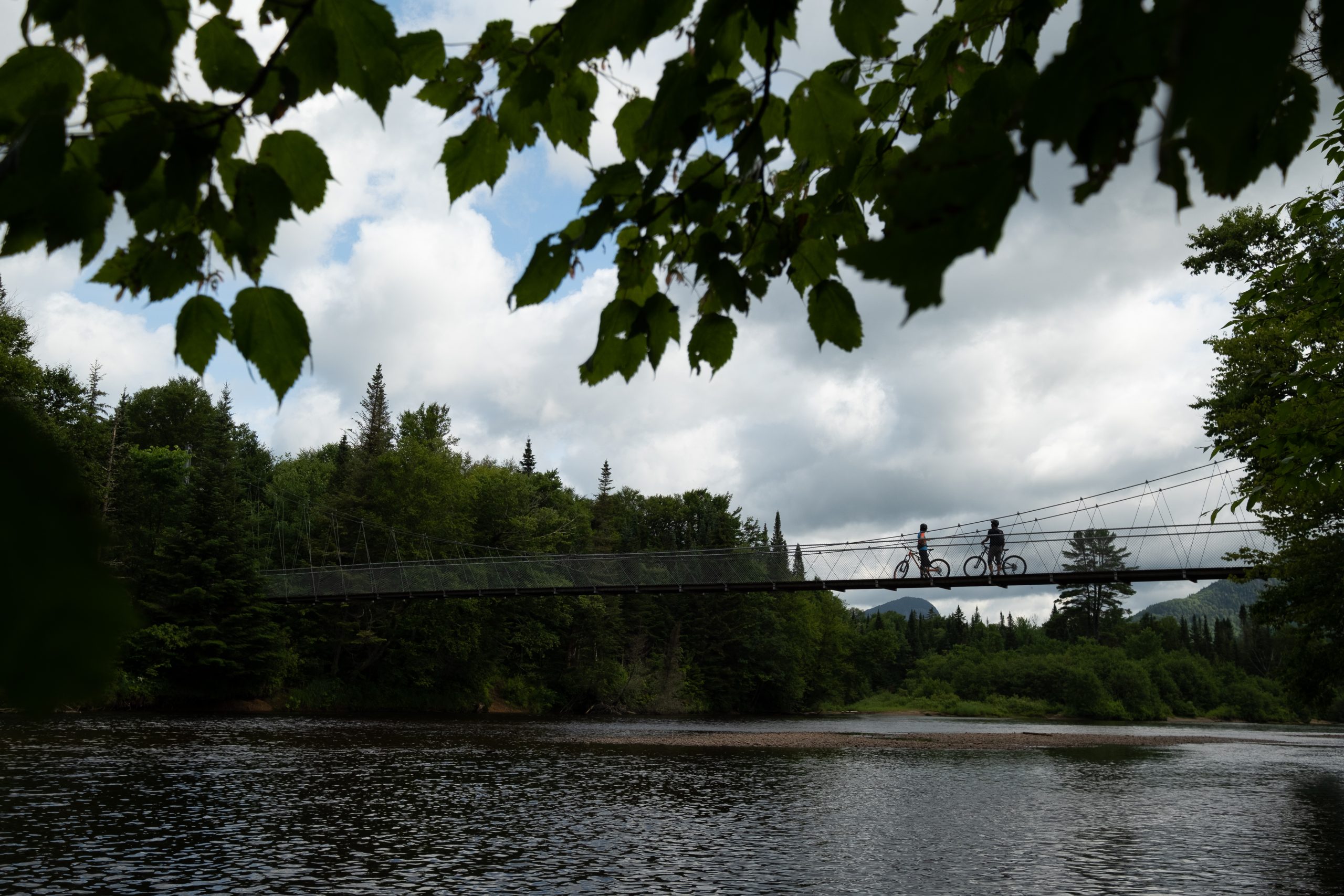 Vue du pont suspendu dans le secteur Shannahan, traversant la rivière, avec deux cyclistes admirant le paysage montagneux qu'offre la nature de la Vallée, lors de leur activité de plein air.