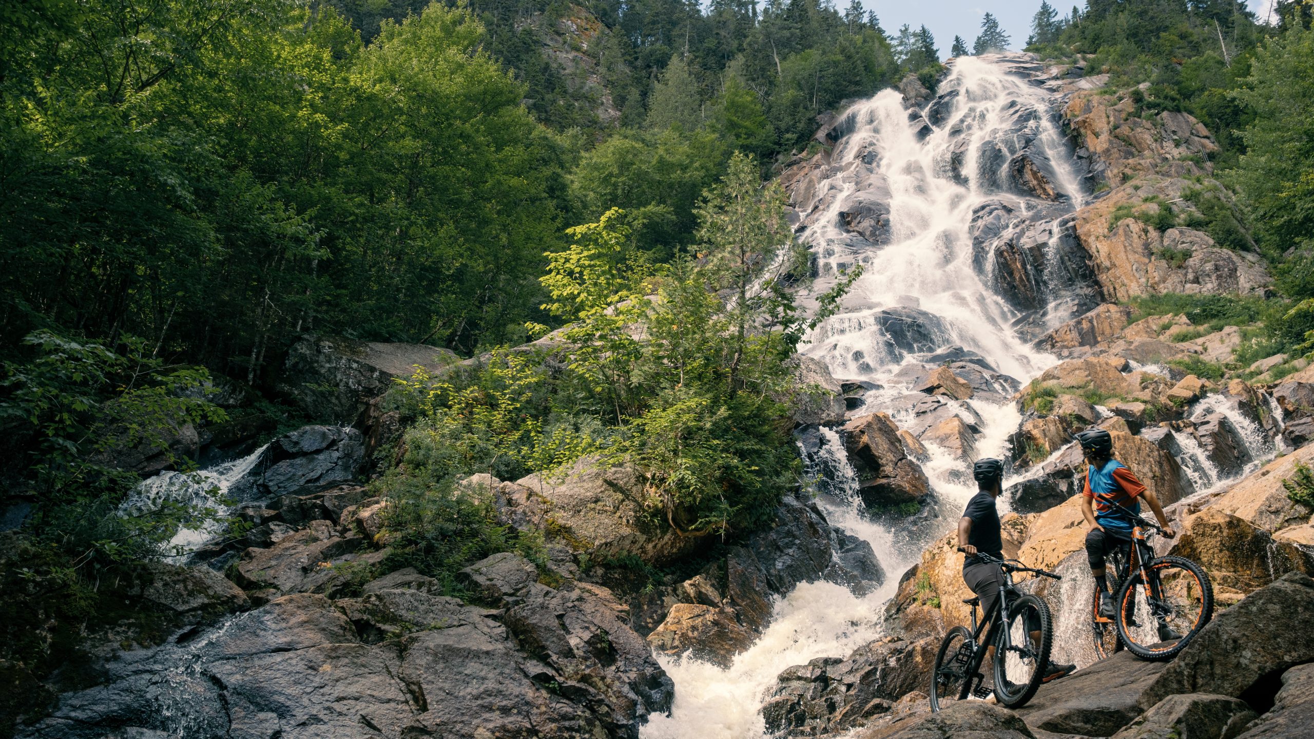 Deux cyclistes prenant une pause en plein air tout en admirant la Chute Delaney, entourée par la nature, l'un des paysages visibles lors de la pratique de sports tels que le vélo de montagne à la Vallée.