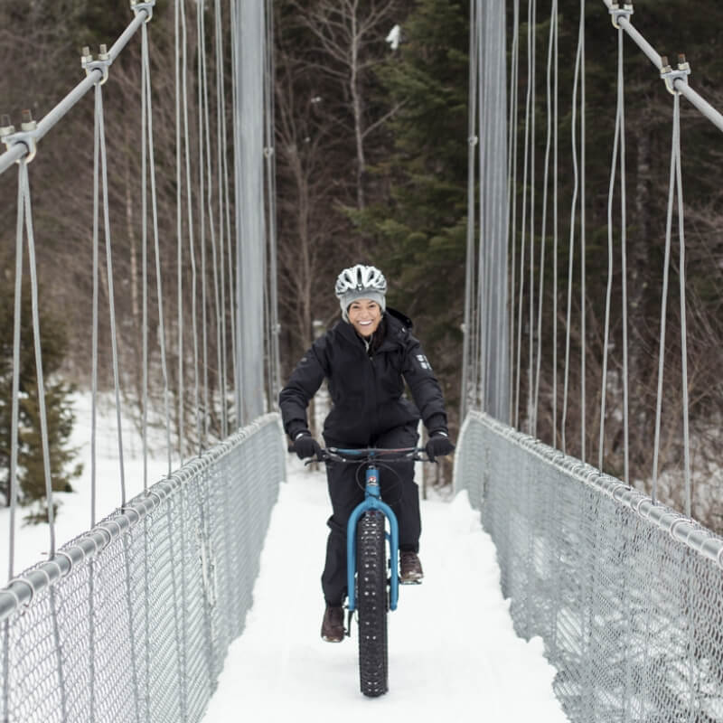 Une femme souriante pratiquant le Fat Bike, soit un sport où l'on peut faire du vélo l'hiver, sur un pont suspendu et enneigé, par l'un des sentiers attitrés de la Vallée.