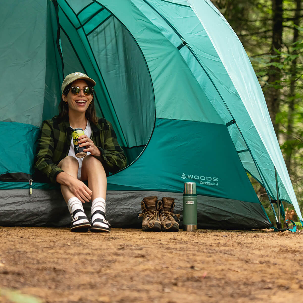 A smiling woman, taking a break on the edge of her camping tent, on one of the sites surrounded by nature and forest at La Vallée.