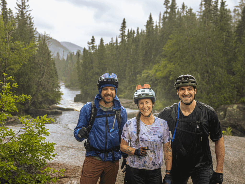 Trois cyclistes souriants profitant d'une sortie sous la pluie près d'une rivière à la Vallée Bras-du-Nord.